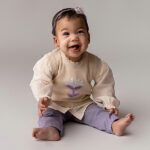 Deze mooie beige spencer met geborduurde bloem komt uit de Simply Nice Girl collectie van Frogs and Dogs. De spencer heeft aan de hals, onderkant en de mouwgaten een gebreid rufflerandje. Op de achterzijde is een sluiting met 3 knoopjes. De geborduurde bloem is in de kleuren gebroken wit & paars. 