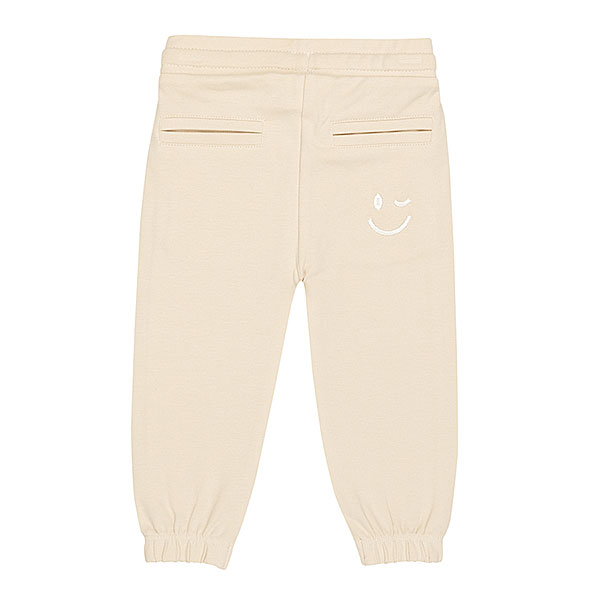 Schattige beige joggingbroek uit de Happy with You collectie van Frogs and Dogs. Het broekje is gemaakt van heerlijk zacht katoen en zit supercomfortabel. De broek heeft twee steekzakjes aan de voorkant en twee nep kontzakjes aan de achterkant. Op een van de achterzakjes zit een geborduurde, blauwe smiley die ondeugend knipoogt — een speels detail!