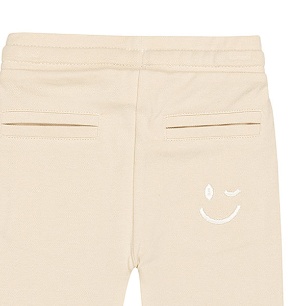 Schattige beige joggingbroek uit de Happy with You collectie van Frogs and Dogs. Het broekje is gemaakt van heerlijk zacht katoen en zit supercomfortabel. De broek heeft twee steekzakjes aan de voorkant en twee nep kontzakjes aan de achterkant. Op een van de achterzakjes zit een geborduurde, blauwe smiley die ondeugend knipoogt — een speels detail!